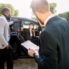 Young business couple, African man and Caucasian woman, looking for car to buy, woman sitting n the car, man standing and smiling. Back view of sales dealer holding the contract.